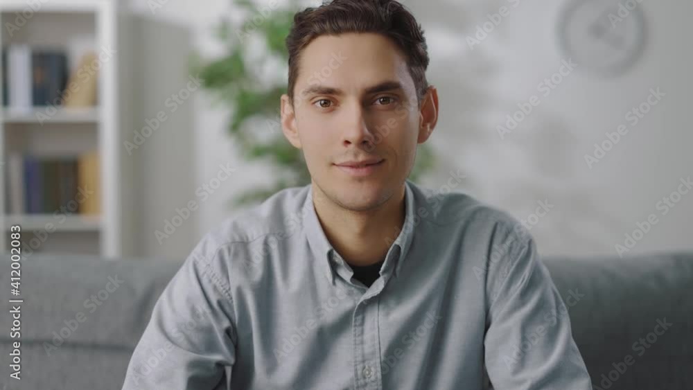 Portrait of relaxed young man in casual shirt and pants sitting on grey couch. Attractive guy with dark hair spending free time at home. Portrait of relaxed young man in casual shirt and pants sitting on grey couch. Attractive guy with dark hair spending free time at home.