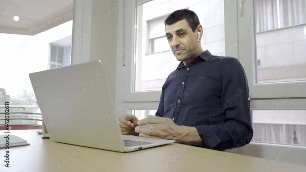 middle-aged dark-haired man in a dark blue shirt teleworking with a laptop, a notebook and a coffee on a wooden table, on call. businessman working on his computer with wireless headphones. video call middle-aged dark-haired man in a dark blue shirt teleworking with a laptop, a notebook and a coffee on a wooden table, on call. businessman working on his computer with wireless headphones. video call