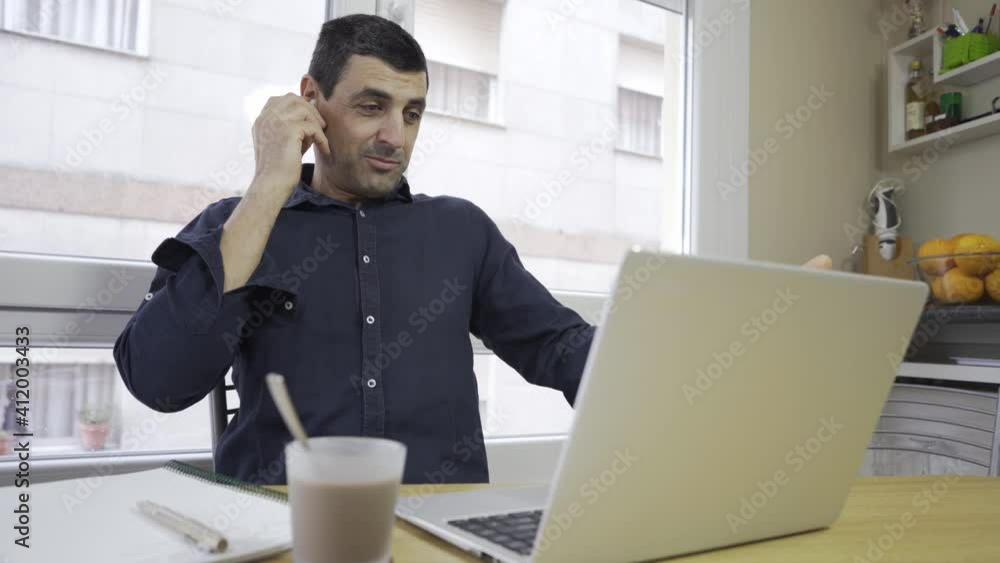 middle-aged dark-haired man in a dark blue shirt teleworking with a laptop, a notebook and a coffee on a wooden table, on call. businessman working on his computer with wireless headphones. video call middle-aged dark-haired man in a dark blue shirt teleworking with a laptop, a notebook and a coffee on a wooden table, on call. businessman working on his computer with wireless headphones. video call