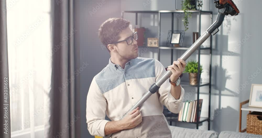Close up portrait of Caucasian young happy handsome man in glasses standing in living room playing vacuum cleaner like guitar, having fun while cleaning apartment, leisure, clean up concept Close up portrait of Caucasian young happy handsome man in glasses standing in living room playing vacuum cleaner like guitar, having fun while cleaning apartment, leisure, clean up concept