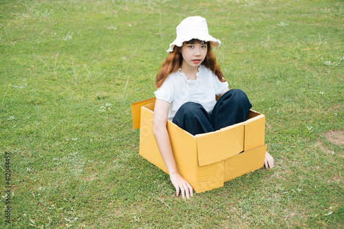 Playful young asian thai woman hipster curly hairstyle on white shirt and hat sitting inside cardboard box on green meadow field in nature. © THESHOTS.CO