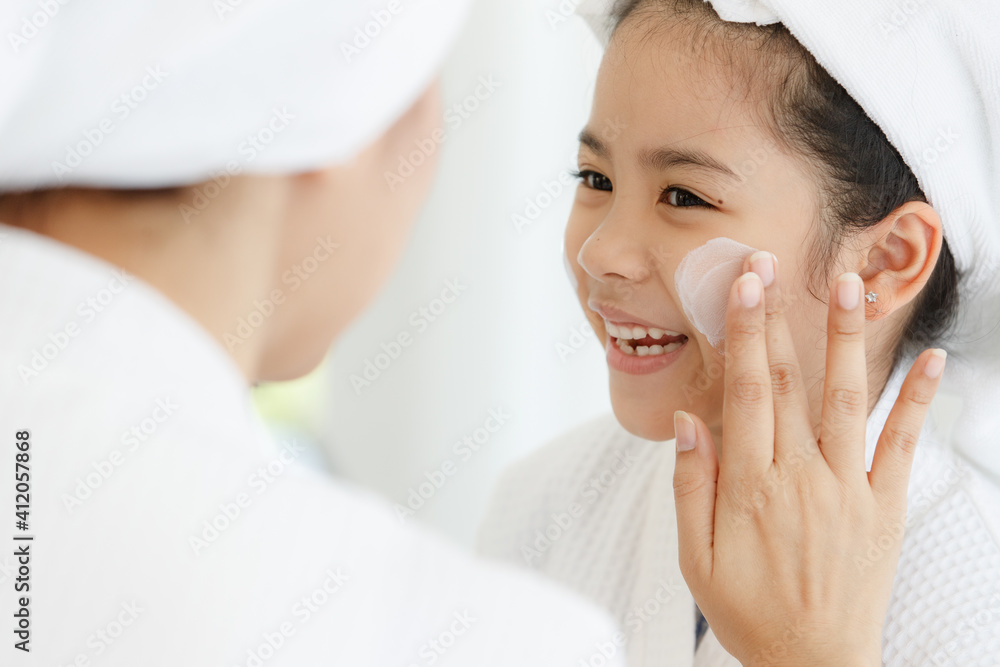 Mother adding treatment cream on the cheek to young and cute Asian girl with spa dress and head covered with a white towel Mother adding treatment cream on the cheek to young and cute Asian girl with spa dress and head covered with a white towel