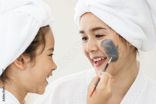 A cute Asian girl with a spa dress and head covered with a white towel using a brush to paint the treatment mud on her mother's cheek. Idea for happiness and relationship of a family