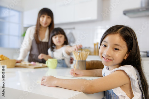 A cute little 7 years old Asian girl sitting and doing homework in kitchen while her mother teaching her younger sister how to make food and cooking in blur background