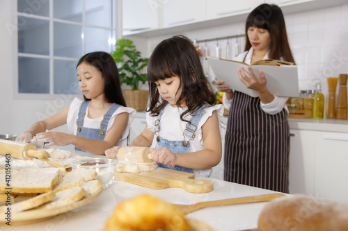 Family group photo of 30s mom teaching and educate two little girls, 3 years and 7 years old, how to make bread and bakery. Mother opening a cookbook to read and find the formula of food cooking