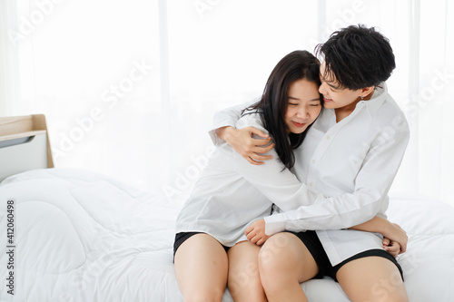 LGBTQ couple lovers, a handsome girl as man or butch hold and cuddle femme girl with tender love, spending and sharing loving time in white bedroom with fun, warmth, and happiness