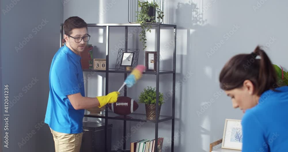 Rear of Caucasian pretty woman talks to handsome young man husband in glasses while he is cleaning furniture wiping dust in living room, family clean up house together, cleaning business concept Rear of Caucasian pretty woman talks to handsome young man husband in glasses while he is cleaning furniture wiping dust in living room, family clean up house together, cleaning business concept