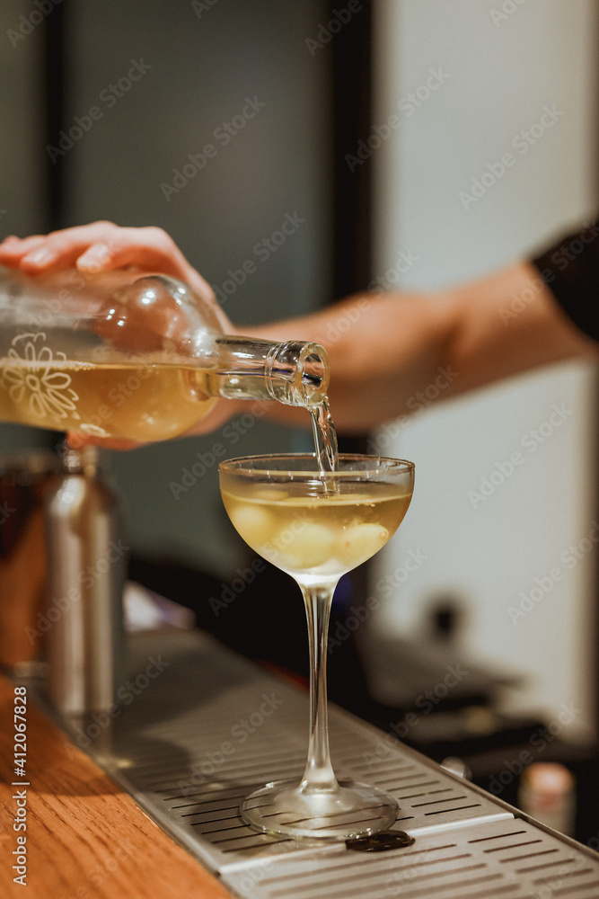 Bartender pouring a drink from a bottle into a coupe cocktail glass. Selective focus. Vertical lifestyle image.