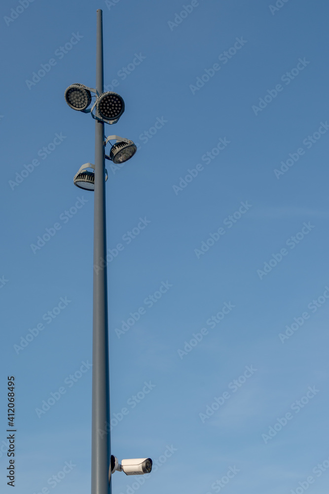 Street metal cone-shaped black pole tapering upward with LED round lights attached to it for night lighting and with a white security camera mounted below, against the blue sky Street metal cone-shaped black pole tapering upward with LED round lights attached to it for night lighting and with a white security camera mounted below, against the blue sky