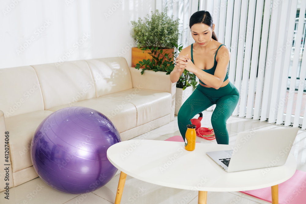 Serious young Asian woman watching workout video on laptop and doing squats at home Serious young Asian woman watching workout video on laptop and doing squats at home
