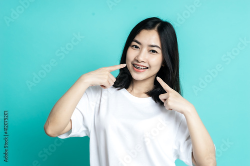 Asian woman wearing dental braces pointing to tooth sample and smiling with her healthy white teeth isolated shot on blue background