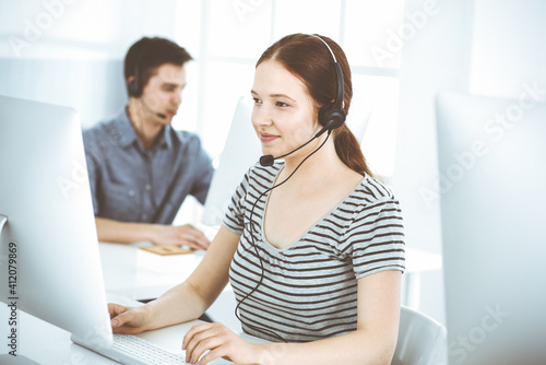 Casual dressed young woman using headset and computer while talking with customers online. Group of operators at work. Call center, business concept