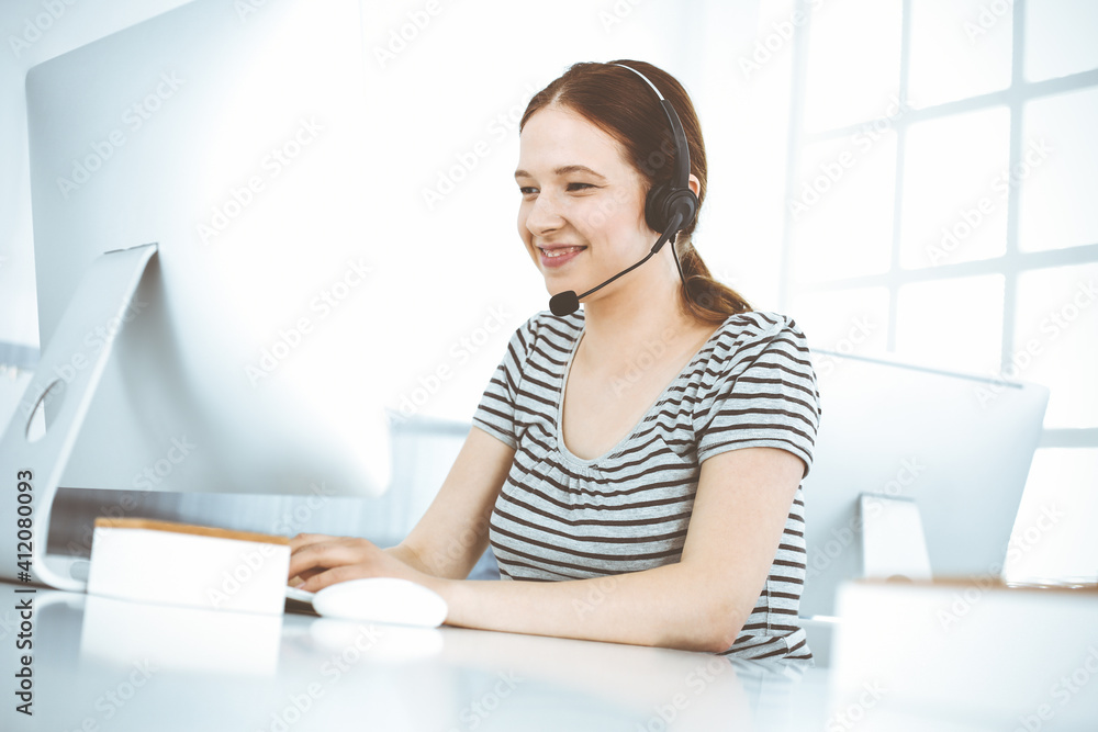 Casual dressed young woman using headset and computer while talking with customers online. Call center, business concept Casual dressed young woman using headset and computer while talking with customers online. Call center, business concept