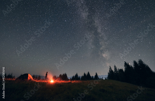 Male hiker standing near campfire and tent under beautiful night sky with stars. Magnificent view of blue starry sky and Milky way under grassy hill. Concept of travelling  hiking and night camping.