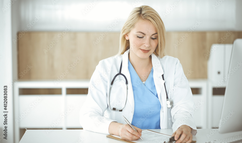 Woman-doctor at work while sitting at the desk in hospital or clinic. Blonde cheerful physician filling up medication history record form Woman-doctor at work while sitting at the desk in hospital or clinic. Blonde cheerful physician filling up medication history record form