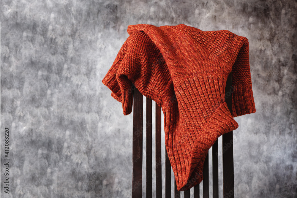 Women's wool knitted terracotta sweater casually thrown over the back of wooden chair against gray wall. Minimal style. Copy space.
