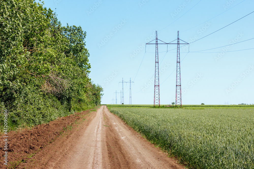 Rural ground road between summer forest and agricultural field with green wheat. Electrical power line towers on farmlands