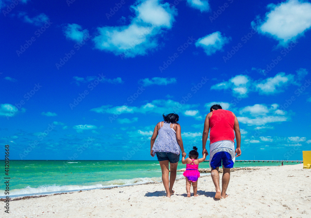 Familia jóven caminando sobre la arena a la orilla de la playa. Padres tomando de la mano a su bebé cerca del mar Familia jóven caminando sobre la arena a la orilla de la playa. Padres tomando de la mano a su bebé cerca del mar