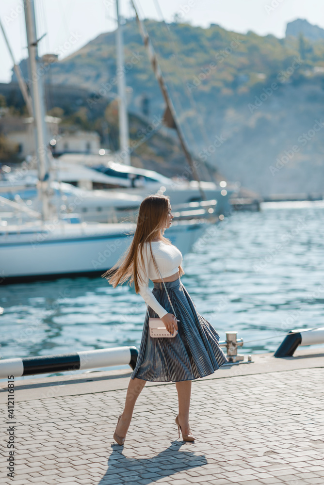 An attractive young fashionable woman dressed in a stylish spring-summer outfit walks along the embankment among the yachts at a luxury resort. Portrait of a beautiful cute girl on a sunny day.