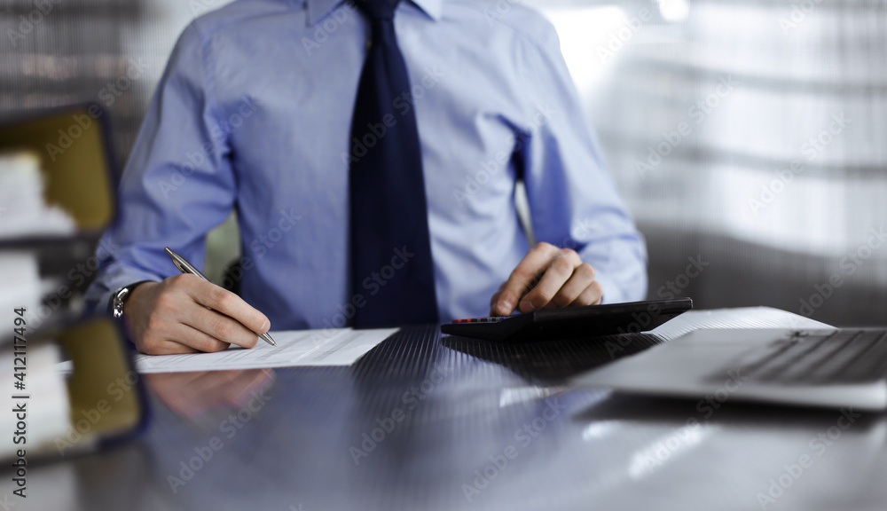 Unknown man accountant in blue shirt use calculator and computer with holding pen on while staying at home during covid pandemic. Taxes and audit concept Unknown man accountant in blue shirt use calculator and computer with holding pen on while staying at home during covid pandemic. Taxes and audit concept