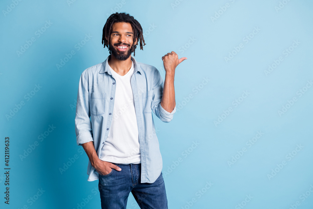 Portrait of positive handsome dark skin guy look indicate thumb empty space isolated on blue color background Portrait of positive handsome dark skin guy look indicate thumb empty space isolated on blue color background