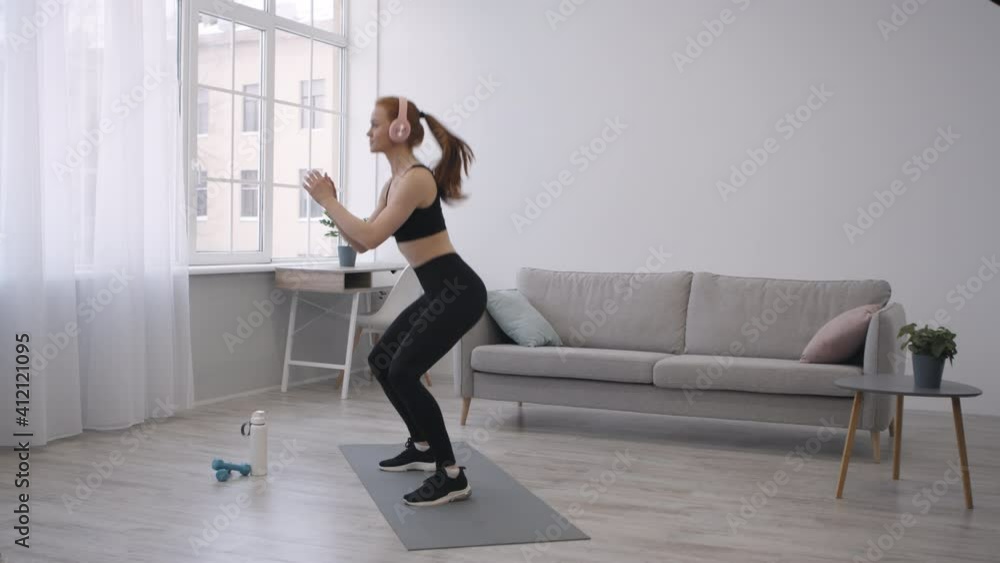 Athletic Woman Doing Jump Squats Exercising Wearing Headphones At Home Athletic Woman Doing Jump Squats Exercising Wearing Headphones At Home