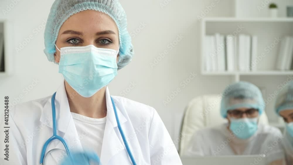 Portrait of female doctor in medical gloves, white coat looks at camera and corrects protective mask on background of colleagues at computer in clinic. Work of doctors during coronavirus pandemic Portrait of female doctor in medical gloves, white coat looks at camera and corrects protective mask on background of colleagues at computer in clinic. Work of doctors during coronavirus pandemic