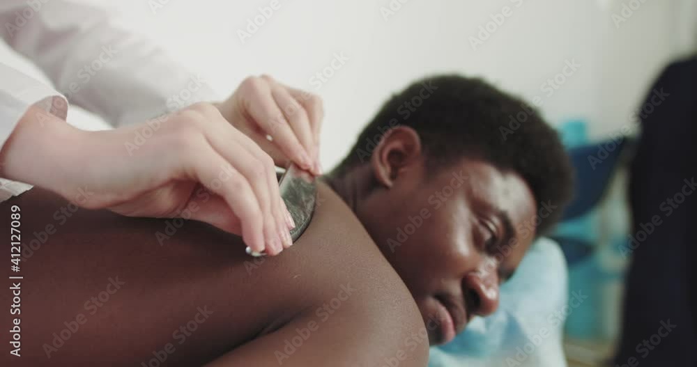 Back Massage Special Device. Young Caucasian Female Physiotherapist. Physiotherapist Conducts Massage Therapy, to African Young Man to Restore Muscle Strength, at the Modern Rehabilitation Clinic. Back Massage Special Device. Young Caucasian Female Physiotherapist. Physiotherapist Conducts Massage Therapy, to African Young Man to Restore Muscle Strength, at the Modern Rehabilitation Clinic.