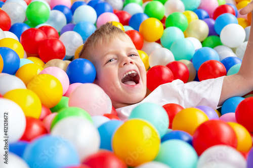 Blonde little boy lying on multi coloured plastic balls in big dry paddling pool in playing centre. Smiling at camera. Portrait close up. Having fun in playroom. Leisure Activity.