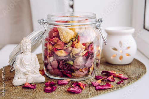 Using dry rose petals to make rose potpourri which is great for home scent. Mason jar filled with dry rose petals and puds with meditating Buddha figurine and aroma lamp with candle next to it.