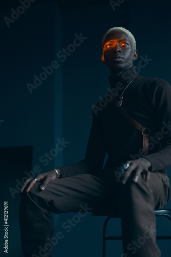 dark-skinned handsome guy with white hair and blue eyes in brown clothes sitting on a chair in a dark studio with a serious expression, where on the eyes shine an orange ray