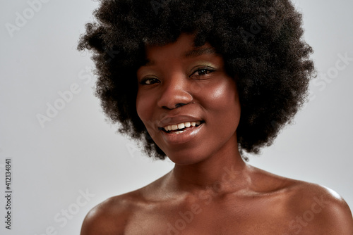 Close up portrait of cheerful african american young woman with afro hair and perfect smooth glowing skin smiling at camera while posing isolated over gray background