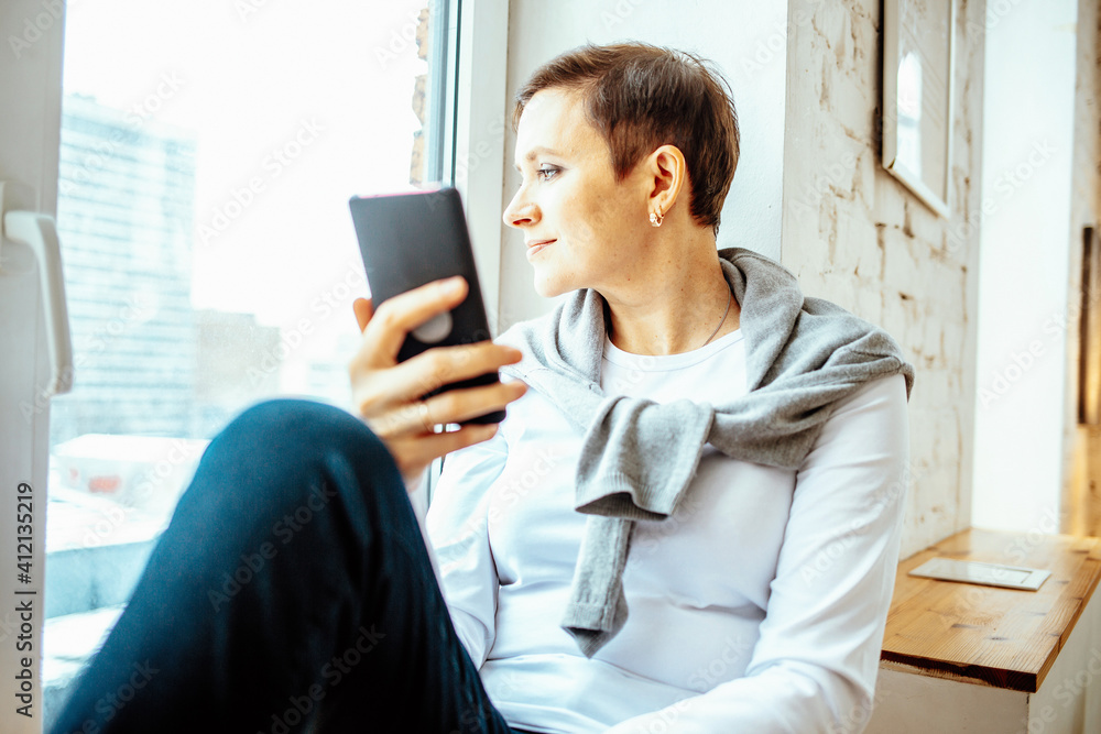 Woman sitting on windowsill with smartphone, working remotely during quarantine Woman sitting on windowsill with smartphone, working remotely during quarantine