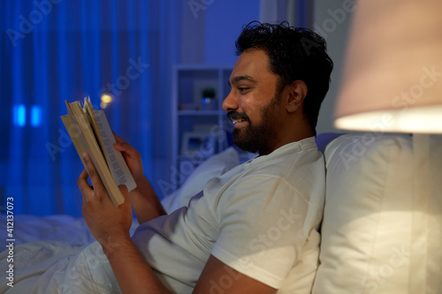 people, bedtime and rest concept - happy smiling indian man reading book in bed at home at night photo