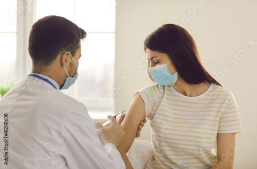 Young female patient looking at man doctor making vaccination against coronavirus infection in her arm in medical clinic office. Vaccine against COVID-19 infection at pandemic times concept