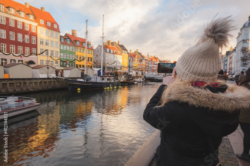 Canvas Print A blonde female tourist in winter photographs colourful cityscape waterfront of Nyhavn Canal (New Harbour), Copenhagen, with her smartphone