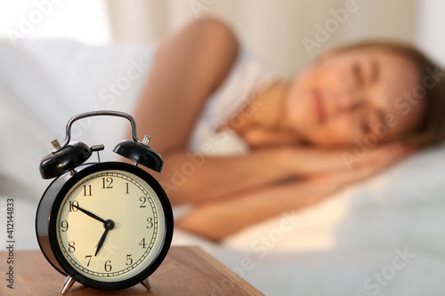 Beautiful young woman sleeping and smiling while lying in bed comfortably and blissfully on the background of alarm clock is going to ring. Sunbeam dawn on her face.