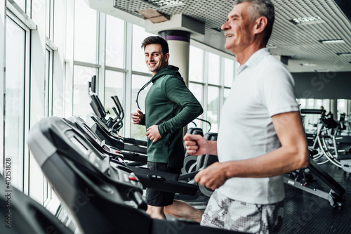 Two people on the treadmill, in the focus of a handsome young coach, he is smiling at the camera. Sports, gym, healthy lifestyle