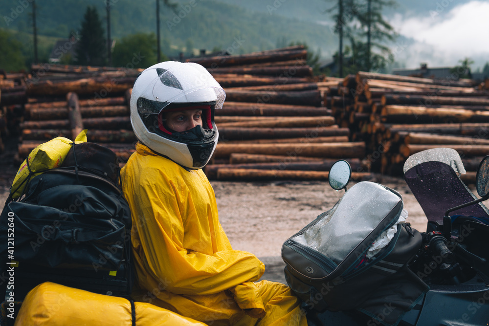 A girl in a raincoat, shoe covers and a helmet is sitting on big adventure motorbike. Vacation and travel. Rain and felling after the storm. Sawmill. Difficult road and biker outfit. Close-up