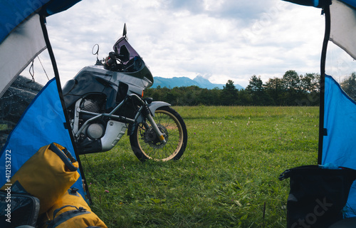 View from camp tent door. Motorcycle adventure bike on the background of green grass, forest and mountains under clouds sky, camping. Copy space for text. Slovenia