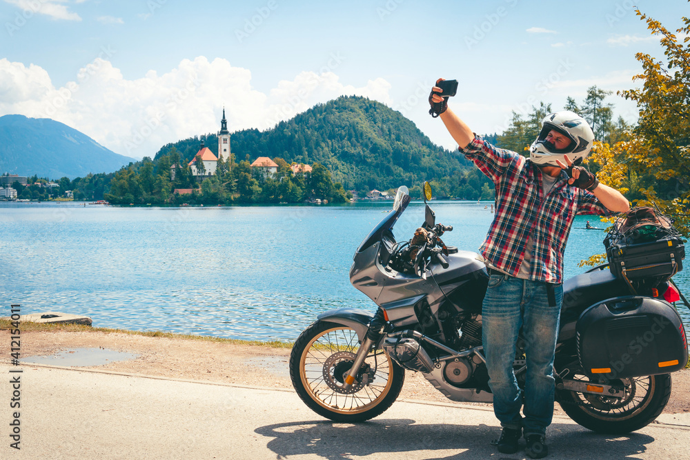 Motorcyclist man takes a selfie on a smartphone. Stands by a motorcycle with bags. Tourism and vacation. Sunny summer day. Bled lake, island, castle and mountains in background, Slovenia, Europe
