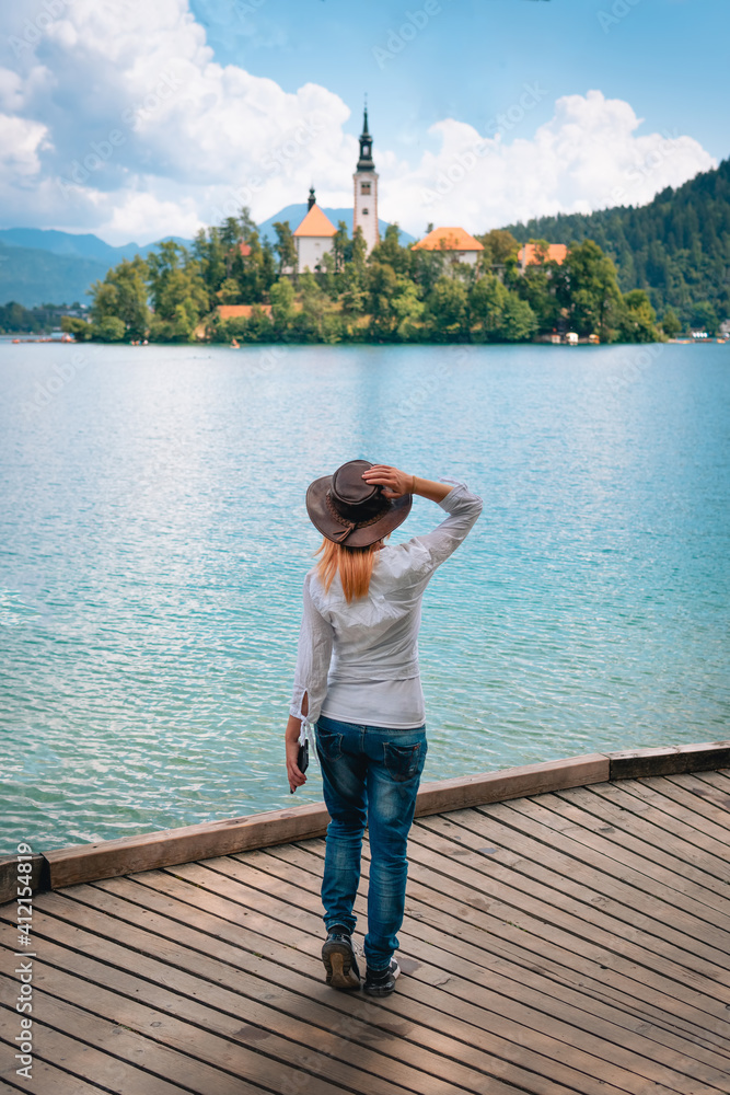 Travel Slovenia, Europe. Back view of young woman posing alone. In a white shirt with sunglasses and a cowboy hat. Bled Lake and Alps Mountain. amazing touristic attractions. Vertical photo