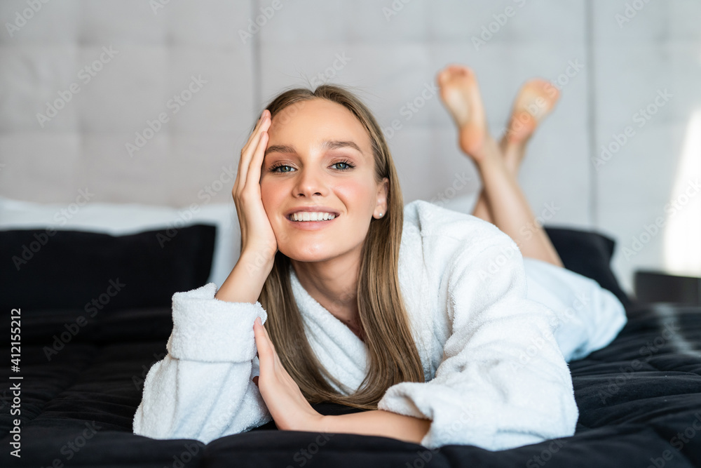 Young beautiful woman in towel and bathrobe sitting on bed at home. Beautiful young woman wearing a white bathrobe, with a towel on her head in bed at home