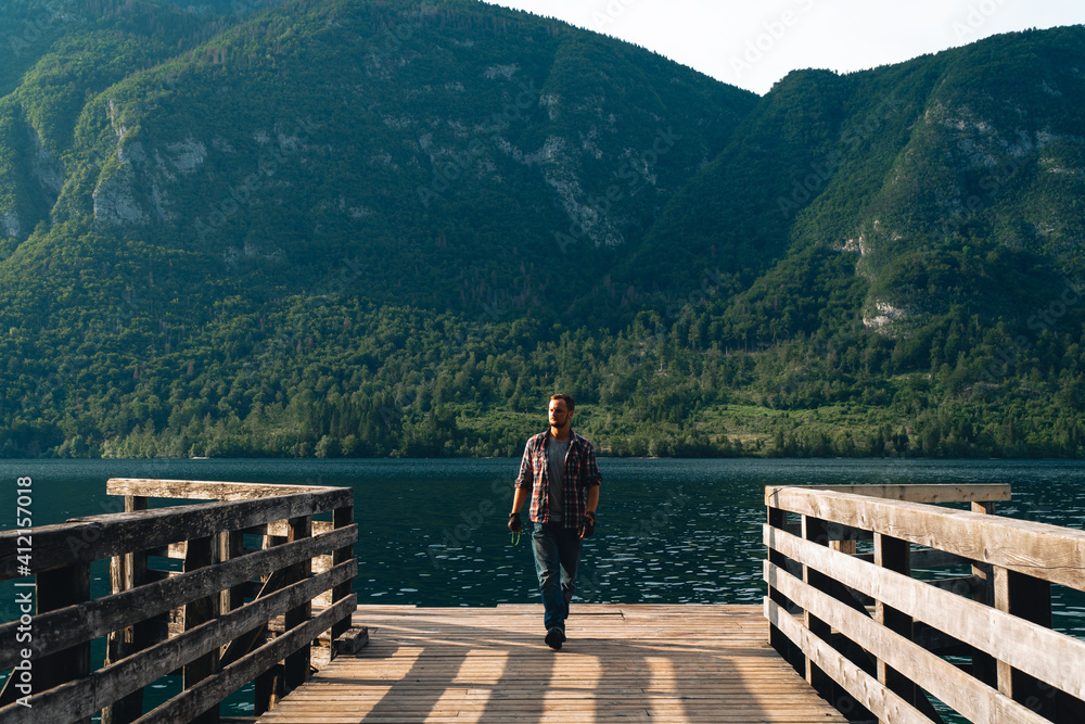 Man alone standing on a wooden pier at a mountains over water. Summer sunset time. Travel and freedom concept 