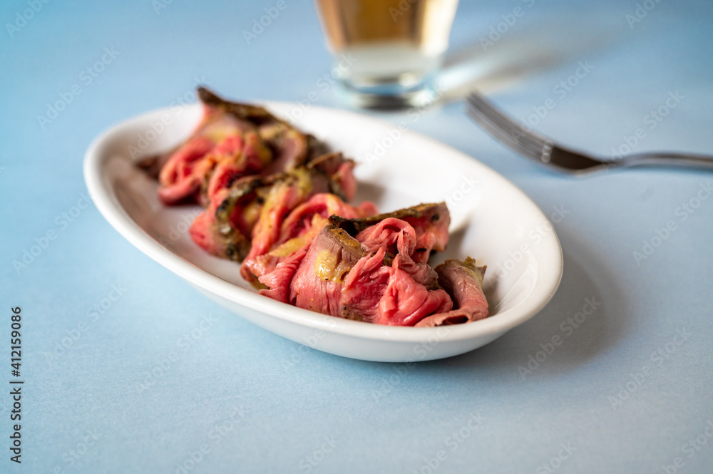 Roast beef with mustard sauce on oval plate, fork and glass of drink on blue background Roast beef with mustard sauce on oval plate, fork and glass of drink on blue background