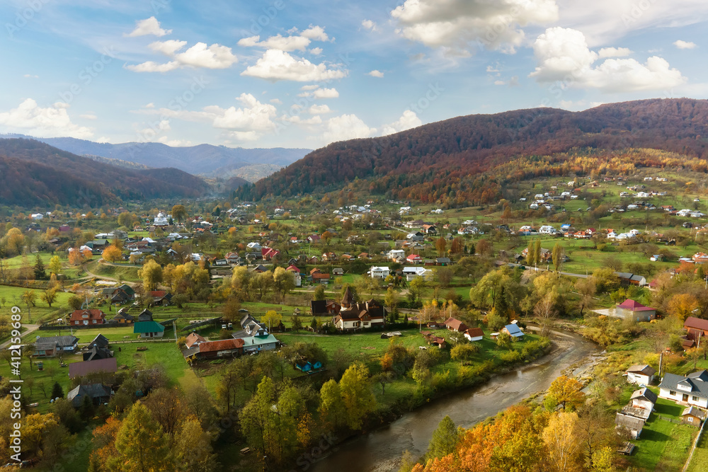 Fototapeta premium Aerial view of a rural village with small houses between autumn mountain hills covered with yellow and green pine trees.