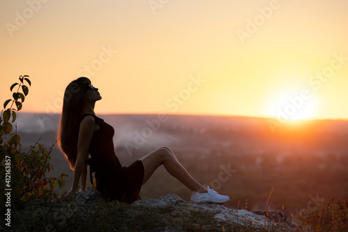 Young elegant woman in black short dress sitting on a rock relaxing outdoors at summer evening. Fashionable female enjoying warm sunset in nature.
