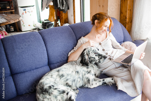 A beautiful young woman is sitting in her house, working on a laptop on the couch with her dog. Stay at home