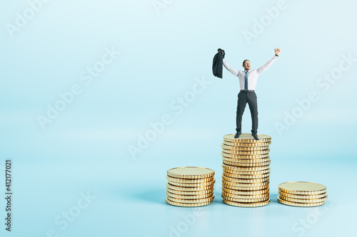 Joyful and successful businessman in a suit standing on a pile of coins at abstract blue background. Earning money concept.