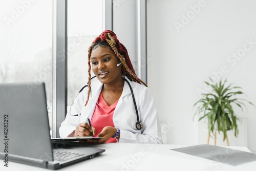 African American woman doctor working at her office online using portable inormation device. Telemedicine services. Primary care consultations, psychotherapy, emergency services. photo